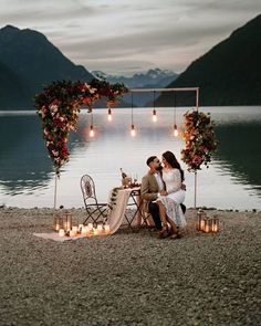 two people sitting at a table with candles on the ground near water and mountains in the background