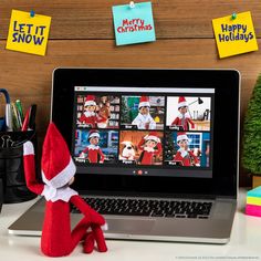 an elf sitting in front of a laptop computer with christmas pictures on the wall behind it