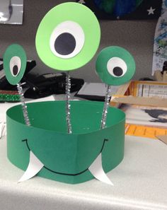 two green paper cups with eyes and sticks sticking out of them on a white table