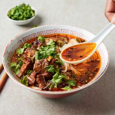 a person holding a spoon in a bowl of soup with meat and vegetables on the side