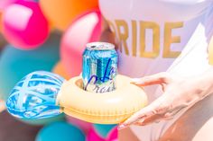 a woman holding up a can of soda in front of colorful balloons and streamers