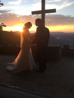 a bride and groom standing in front of a cross with the sun setting behind them