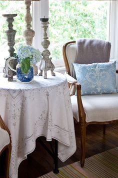 a white table topped with a vase filled with flowers next to a chair and window