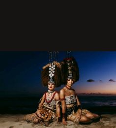 two people sitting on the beach at night with their headdresses in front of them