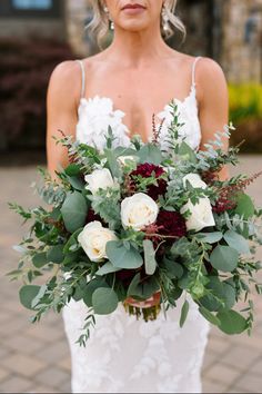 a woman holding a bouquet of flowers and greenery on her wedding day in front of a brick building