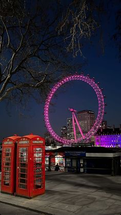 two red telephone booths sitting next to each other near a tree and ferris wheel in the background