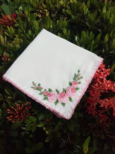 a white handkerchief with pink roses on it sitting in the grass next to some red flowers