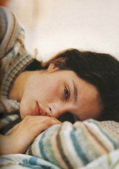 a woman laying in bed under a blanket with her hand on her chin and looking at the camera