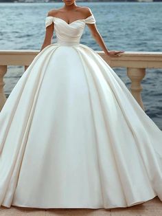 a woman in a white wedding dress standing on a balcony near the water with her hands on her hips