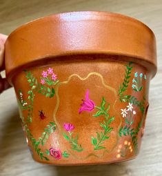 a hand holding a brown pot with flowers painted on the side and green leaves around it