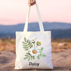 a person holding a white bag with daisies and leaves on it in the sand