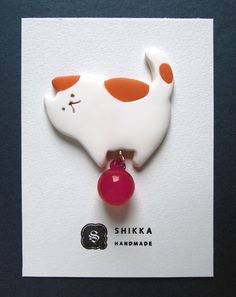 a white and orange cat brooch with a red ball on it's back