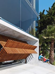 two people sitting on a bench in front of a building with wooden slatted design