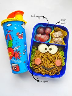 a lunch box with noodles, apples, and an owl face on it next to a plastic cup