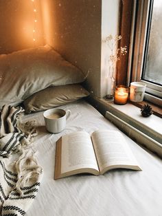 an open book and cup sit on a bed next to a window sill with candles