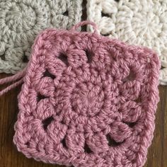 three crocheted squares are sitting on a wooden surface, one is pink and the other is white