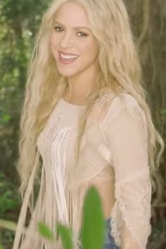 a woman with long blonde hair standing in front of trees and smiling at the camera