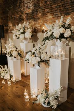 several white vases filled with flowers and candles on a wooden floor in front of a brick wall