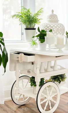 a white cart with plants and candles on it sitting in front of a window next to a potted plant