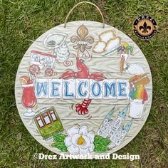 a welcome sign is sitting in the grass with flowers and other things around it that are painted on