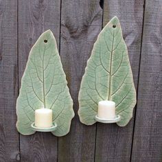 two green leaf shaped candle holders with white candles on them sitting next to a wooden fence