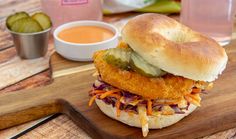 a chicken sandwich with coleslaw and pickles on a cutting board next to dipping sauces