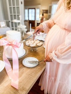 a pregnant woman cutting into a cake with a pink ribbon on the table next to it