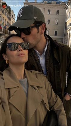 a man kissing a woman on the cheek while she is wearing sunglasses and a trench coat