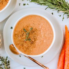 two white bowls filled with soup next to some carrots and sprig of rosemary