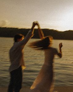 two people are dancing by the water at sunset with their arms in the air and one person is holding his hands up