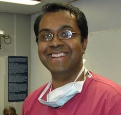 a man with glasses and a pink shirt smiles at the camera while standing in a hospital room