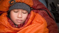 a woman in an orange sleeping bag with her eyes closed and head tucked under the hood