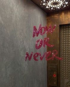 the word now or never is painted on a wall in an elevator with lights above it