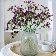 purple flowers are in a glass vase on a plate with a cup and saucer