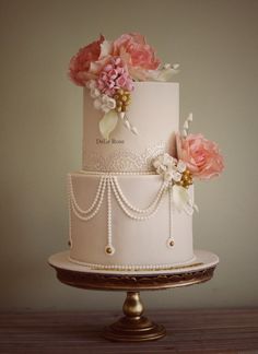 a three tiered wedding cake with pink flowers and pearls on the top, sitting on a wooden table