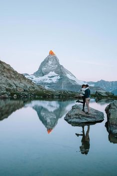 two people standing on top of a rock in front of a body of water with mountains in the background