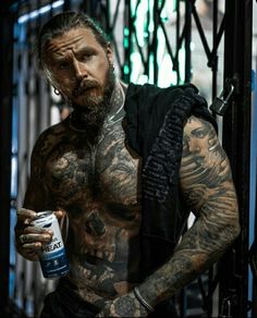 a man with tattoos and piercings holding a can of beer in his hand while standing next to an iron gate