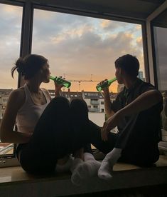 two people are sitting on a window sill drinking beer and looking out at the city