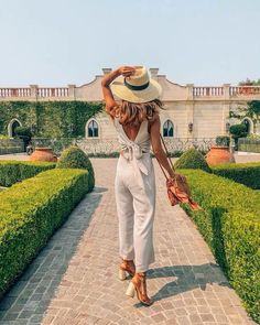 a woman wearing a hat and white jumpsuit walking down a brick walkway in front of hedges