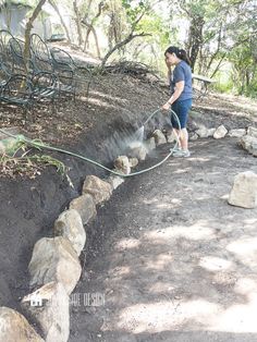 a woman is using a hose to clean the soil in her garden area with rocks