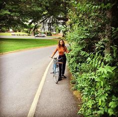 a woman is riding her bike down the street in front of some bushes and trees