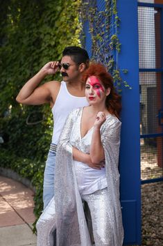 a man standing next to a woman with red makeup on her face and wearing silver pants