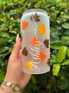 a hand holding up a frosted can with autumn leaves painted on it and the words happy written in white lettering