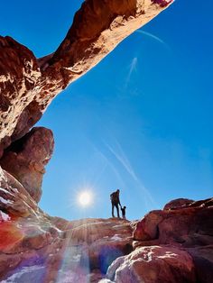 two people standing on top of a rocky mountain under a blue sky with the sun shining