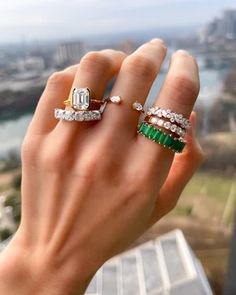 a woman's hand holding three rings with emeralds and diamonds on them in front of a cityscape