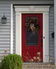 a red front door with a wreath on it and some plants in the foreground