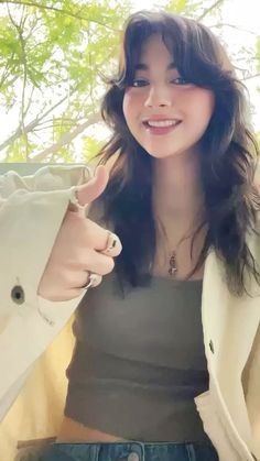 a woman with long dark hair wearing a white jacket and black shirt giving the thumbs up sign