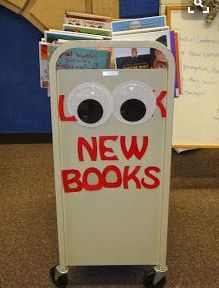 a cardboard box with eyes and the words new books on it sitting in front of a bulletin board