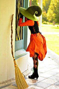 a woman in an orange dress and green hat leaning against a door with a broom