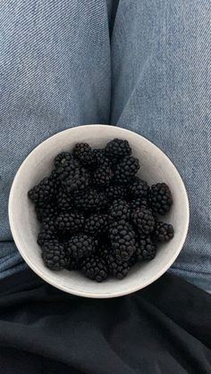 a white bowl filled with blackberries sitting on top of someone's legs in blue jeans